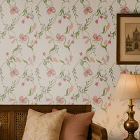 A warm and elegant living room featuring soft pink floral wallpaper with delicate green vines and blossoms. A wooden daybed-style sofa with a cane back and cream upholstery is accented with blush pink and beige cushions. Matching wooden side tables hold a decorative lantern and a table lamp, while a simple coffee table with books and a decorative bowl sits on a natural woven rug. Sunlight streams through a carved wooden window, creating soft shadows and enhancing the room's cozy, traditional ambiance.