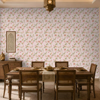 A warm and inviting dining room featuring soft pink floral wallpaper with delicate green vines and blossoms. A solid wood dining table with six cane-back chairs is set with neutral-toned dinnerware and a beige table runner. To the left, a wooden sideboard holds a decorative lantern and stacked books beneath a framed carved wall panel. A hanging lantern-style ceiling light, carved wooden door, sheer curtains, and natural sunlight create a cozy, elegant ambiance with a blend of traditions 
