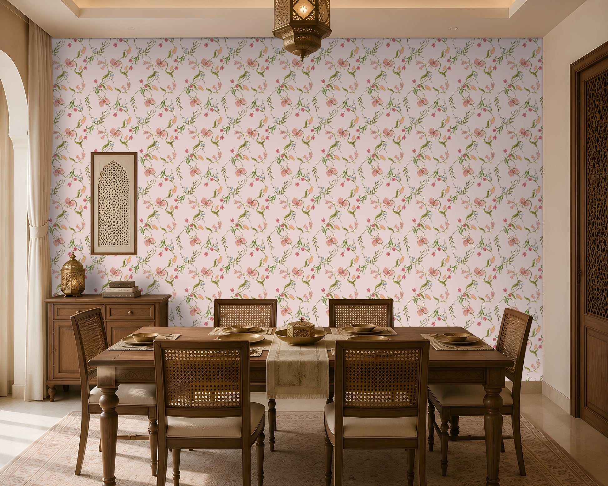 A warm and inviting dining room featuring soft pink floral wallpaper with delicate green vines and blossoms. A solid wood dining table with six cane-back chairs is set with neutral-toned dinnerware and a beige table runner. To the left, a wooden sideboard holds a decorative lantern and stacked books beneath a framed carved wall panel. A hanging lantern-style ceiling light, carved wooden door, sheer curtains, and natural sunlight create a cozy, elegant ambiance with a blend of traditions 