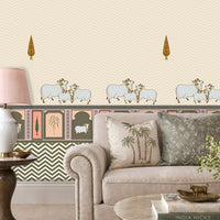 An elegant living room with off-white patterned wallpaper featuring subtle chevron textures, small tree motifs, and a decorative border with animal illustrations. In front is a beige upholstered sofa with cushions, including palm tree and geometric designs. A wooden side table holds a white lamp with a soft pink shade, while a round coffee table displays books, a small brass animal figurine, and a vase of flowers. 
