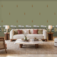 Green-toned room with patterned wallpaper featuring small decorative motifs and a horizontal border with sheep illustrations. A white vintage-style desk with curved legs sits against the wall, paired with a cushioned stool. On the desk is a patterned lamp and an open book. To the left, tall glass-paneled doors let in natural light, and the floor is tiled in a light color with small black diamond accents.