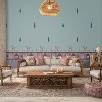 An elegantly decorated living room featuring a large accent wall with light blue wallpaper. The wallpaper is adorned with a pattern of small, stylized teal cypress trees and a decorative border along the bottom showing white cows and traditional arch motifs in purple and pink.