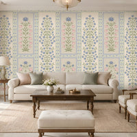 Elegant contemporary living room featuring blue, pastel pink, and muted sage green floral wallpaper with intricate botanical motifs and decorative panel borders on a warm ivory background. The heritage-inspired wallpaper creates a sophisticated focal wall, complemented by a cream upholstered sofa, matching accent armchairs, neutral cushions, a dark wooden coffee table, and a textured beige rug.
