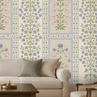 Elegant contemporary living room featuring blue, pastel pink, and muted sage green floral wallpaper with intricate botanical motifs and decorative panel borders on a warm ivory background. The heritage-inspired wallpaper creates a sophisticated focal wall, complemented by a cream upholstered sofa, matching accent armchairs, neutral cushions, a dark wooden coffee table, and a textured beige rug.