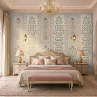 Luxurious heritage-style bedroom featuring pastel purple, dusty pink, and muted sage green floral wallpaper with intricate botanical motifs and decorative panel borders on a soft ivory background. The elegant wallpaper complements a cream upholstered bed with a carved cane headboard, blush pink cushions and bedding, a matching pink upholstered bench, and ornate ivory bedside tables.
