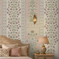 Luxurious heritage-style bedroom featuring pastel purple, dusty pink, and muted sage green floral wallpaper with intricate botanical motifs and decorative panel borders on a soft ivory background. The elegant wallpaper complements a cream upholstered bed with a carved cane headboard, blush pink cushions and bedding, a matching pink upholstered bench, and ornate ivory bedside tables.