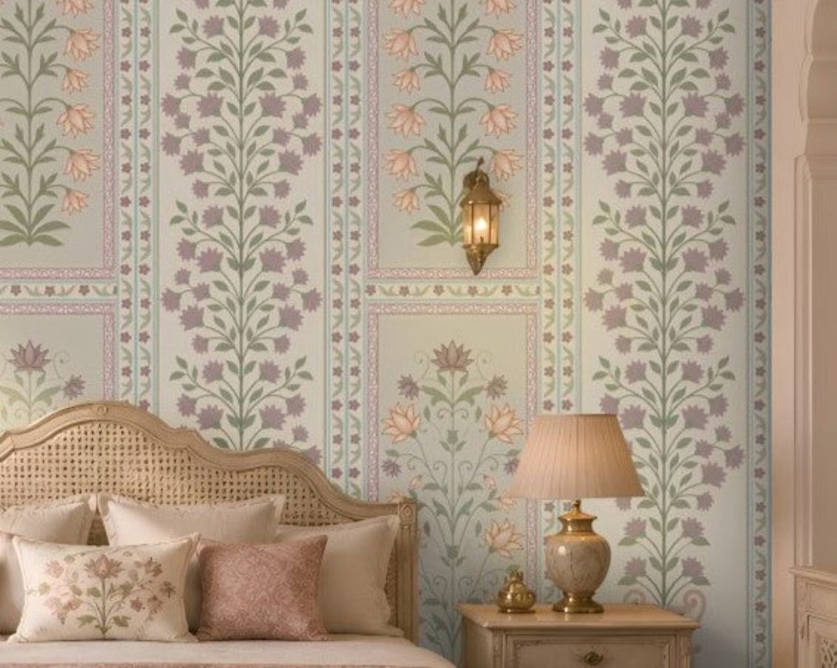 Luxurious heritage-style bedroom featuring pastel purple, dusty pink, and muted sage green floral wallpaper with intricate botanical motifs and decorative panel borders on a soft ivory background. The elegant wallpaper complements a cream upholstered bed with a carved cane headboard, blush pink cushions and bedding, a matching pink upholstered bench, and ornate ivory bedside tables.
