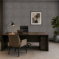 A modern home office setup featuring a dark wood desk with a beige upholstered chair, set against a textured gray wall with a framed abstract print. A black office chair, desk lamp, and a potted green plant are arranged around the desk, with a built-in wooden shelving unit on the left and a large window on the right.