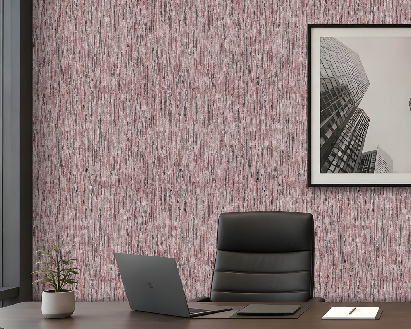 A styled office interior featuring textured wallpaper with a vertical brushstroke pattern in dusty pink, mauve, and gray. A dark gray leather office chair sits on a dark wood desk next to an open laptop, with a small potted plant and a framed black-and-white cityscape print on the wall.