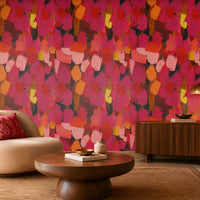 Modern Expression Coral Red abstract wallpaper featuring a bold, artistic brushstroke pattern in vibrant coral red, deep crimson, magenta, dusty pink, fiery orange, golden yellow, and charcoal accents. Displayed on a seamless statement wall in a contemporary living room, this expressive multicoloured wallpaper creates a warm, dramatic, and luxurious atmosphere. The interior features a cream boucle accent chair with a decorative red cushion, a round wooden coffee table, a dark wood fluted sideboard, a brass 