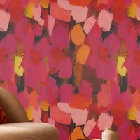 Modern Expression Coral Red abstract wallpaper featuring a bold, artistic brushstroke pattern in vibrant coral red, deep crimson, magenta, dusty pink, fiery orange, golden yellow, and charcoal accents. Displayed on a seamless statement wall in a contemporary living room, this expressive multicoloured wallpaper creates a warm, dramatic, and luxurious atmosphere. The interior features a cream boucle accent chair with a decorative red cushion, a round wooden coffee table, a dark wood fluted sideboard, a brass 