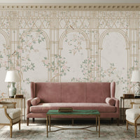 Morning Dew Trellis wallpaper in warm ivory and champagne tones featuring elegant trellis arches, delicate flowering vines, perched birds, butterflies, and soft botanical details. Styled in a sophisticated living room with a blush velvet sofa, classic armchairs, decorative table lamps, a green marble coffee table, and ornate architectural moldings, the mural creates a refined garden-inspired backdrop with timeless elegance and luxurious charm.