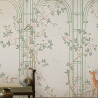 Morning Dew Trellis wallpaper in soft sage green featuring elegant trellis arches, delicate flowering vines, perched birds, graceful butterflies, and botanical details. Displayed in a sophisticated living room with a coffered wood ceiling, ornate chandelier, plush seating, brass accents, and warm wood flooring, the mural creates a serene garden-inspired backdrop that blends timeless elegance with luxurious interior design.