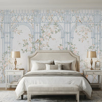 Morning Dew Trellis wallpaper in powder blue showcasing elegant architectural arches and trellis detailing intertwined with delicate flowering vines, soft pastel foliage, perched birds, and subtle botanical motifs. Displayed in a sophisticated bedroom with a cream upholstered bed, matching bedside tables, warm wood flooring, and refined décor, creating a serene, timeless, garden-inspired atmosphere.