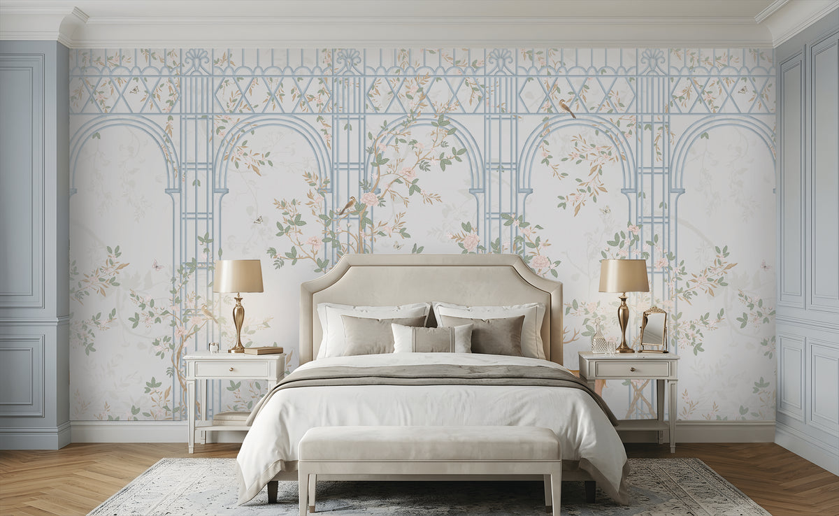 Morning Dew Trellis wallpaper in powder blue showcasing elegant architectural arches and trellis detailing intertwined with delicate flowering vines, soft pastel foliage, perched birds, and subtle botanical motifs. Displayed in a sophisticated bedroom with a cream upholstered bed, matching bedside tables, warm wood flooring, and refined décor, creating a serene, timeless, garden-inspired atmosphere.
