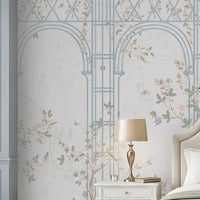 Morning Dew Trellis wallpaper in powder blue showcasing elegant architectural arches and trellis detailing intertwined with delicate flowering vines, soft pastel foliage, perched birds, and subtle botanical motifs. Displayed in a sophisticated bedroom with a cream upholstered bed, matching bedside tables, warm wood flooring, and refined décor, creating a serene, timeless, garden-inspired atmosphere. 