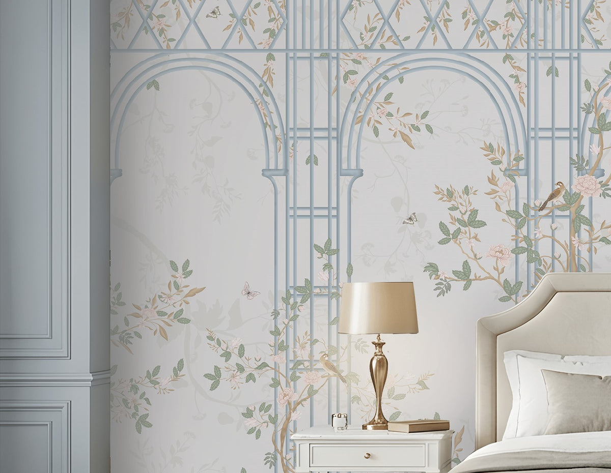 Morning Dew Trellis wallpaper in powder blue showcasing elegant architectural arches and trellis detailing intertwined with delicate flowering vines, soft pastel foliage, perched birds, and subtle botanical motifs. Displayed in a sophisticated bedroom with a cream upholstered bed, matching bedside tables, warm wood flooring, and refined décor, creating a serene, timeless, garden-inspired atmosphere. 