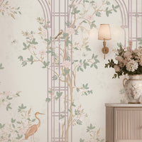 Morning Dew Trellis wallpaper in soft blush pink featuring elegant trellis arches, delicate flowering branches, perched birds, butterflies, and graceful botanical details. Styled in a sophisticated entryway with a curved gold-framed mirror, floral chandelier, fluted console table, decorative ceramic vases, and warm ambient lighting, the mural creates a serene garden-inspired backdrop that blends timeless elegance with romantic luxury.