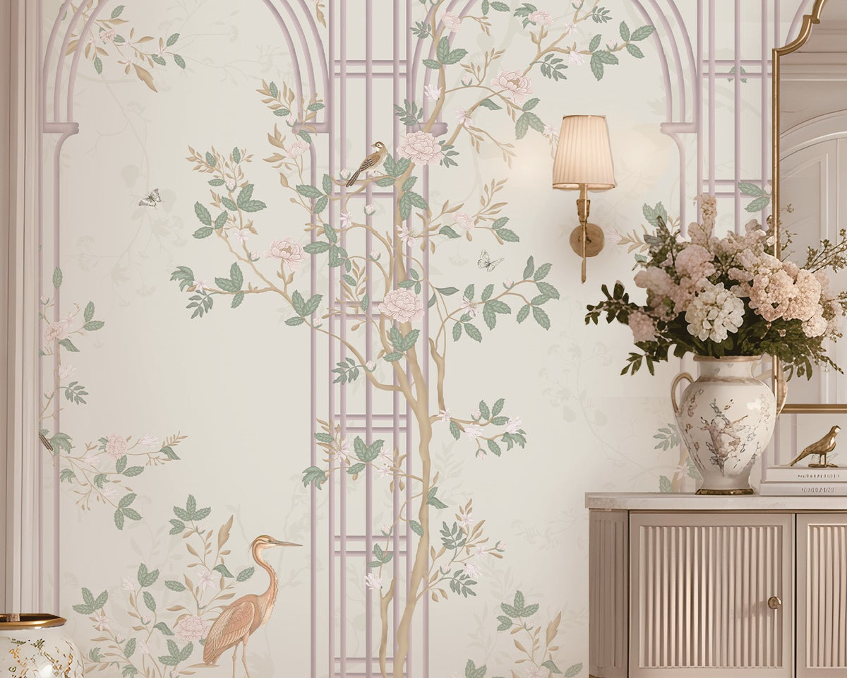 Morning Dew Trellis wallpaper in soft blush pink featuring elegant trellis arches, delicate flowering branches, perched birds, butterflies, and graceful botanical details. Styled in a sophisticated entryway with a curved gold-framed mirror, floral chandelier, fluted console table, decorative ceramic vases, and warm ambient lighting, the mural creates a serene garden-inspired backdrop that blends timeless elegance with romantic luxury.