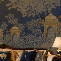 An ornate interior scene featuring a dark navy blue wallpaper with a gold and beige toile-style pattern of domes, trees, and foliage, behind a carved sofa with decorative pillows and a lit table lamp.