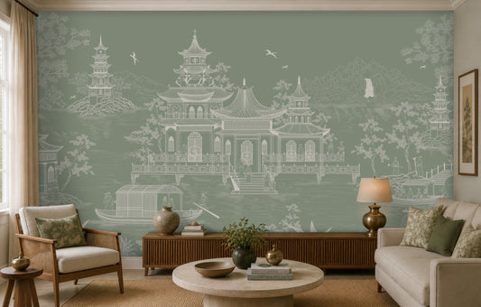 A styled living room featuring a large wall mural with a pale sage green background and a white line-art illustration of traditional East Asian pagodas, boats, and birds. The room is furnished with a cream sofa, a wooden sideboard, a round coffee table, and matching green accent pillows.