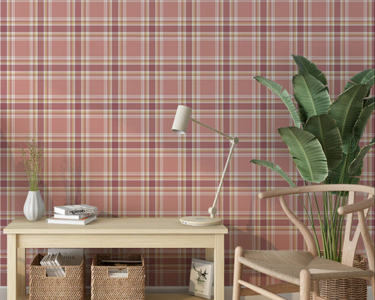 A brightly lit interior space featuring a blushed brick-red and cream plaid wallpaper accent wall. In the center sits a low, light wood console table holding a small white fluted vase with sprigs, a short stack of books, and a sleek cream desk lamp. Underneath the table, two woven wicker baskets store books and magazines. To the left, a woven basket holds tall, decorative dried fan palm leaves, while a modern light wood wishbone chair rests on the right next to a large potted banana leaf plant.
