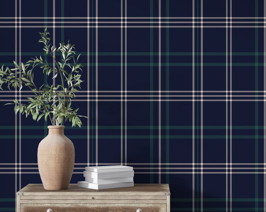 An interior setting highlights a dark navy blue wallpaper with a classic green and white plaid pattern.
In the foreground, a rustic light wood chest or table sits with subtle metal rivets along its top edge.
A large, textured terracotta vase with a rounded body rests on the left side of the wooden surface.
The vase is filled with leafy olive branches extending upwards, adding a vibrant touch of natural green.