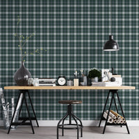 A modern workspace or studio setting featuring a wall covered in a classic plaid wallpaper pattern with a deep juniper green background, crisscrossed by white and muted grey stripes. In the foreground, a light-colored wooden desk is organized with various items, including stacks of books, a black vintage-style twin-bell alarm clock, small jars, paintbrushes in containers, and a small potted green plant. A matte black dome pendant light hangs over the right side of the desk, illuminating the workspace.