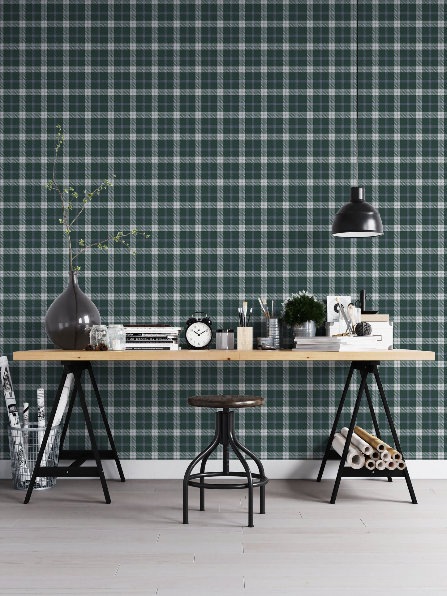 A modern workspace or studio setting featuring a wall covered in a classic plaid wallpaper pattern with a deep juniper green background, crisscrossed by white and muted grey stripes. In the foreground, a light-colored wooden desk is organized with various items, including stacks of books, a black vintage-style twin-bell alarm clock, small jars, paintbrushes in containers, and a small potted green plant. A matte black dome pendant light hangs over the right side of the desk, illuminating the workspace.