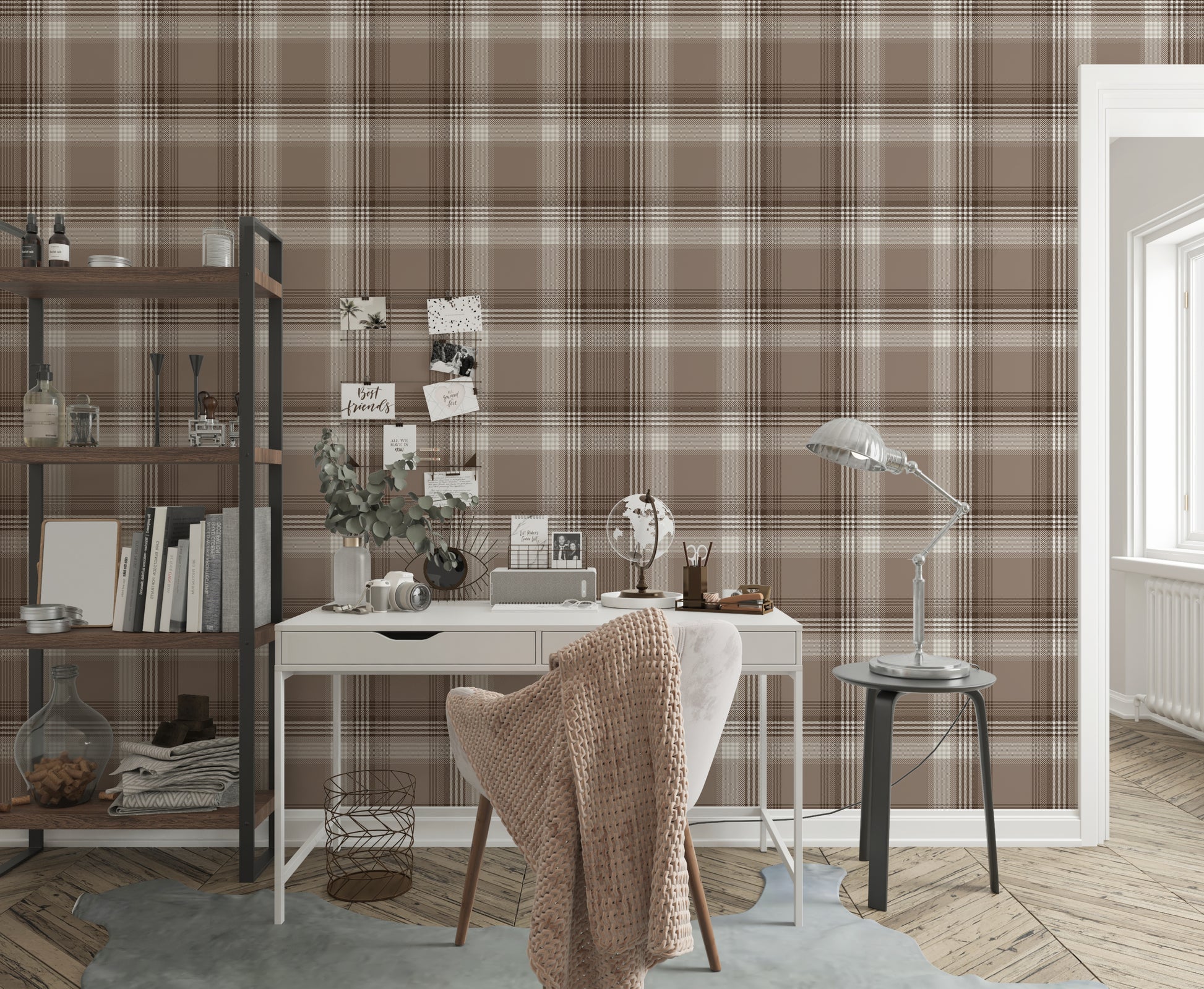A styled home office area featuring a prominent faded mocha brown and white plaid wallpaper. In the center, a white writing desk is paired with a matching white chair draped with a chunky beige knit blanket. The desk is organized with a small globe, a desk organizer, a camera, and a vase with eucalyptus branches. To the left stands a tall industrial-style wooden bookshelf with a black metal frame, holding books and decor. To the right, a silver metal task lamp rests on a small black side table near an open 
