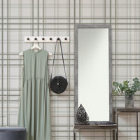 A warm interior setup features a soft faded olive green and grey plaid wallpaper.
The detailed design shows thin dark lines intersecting to form clean grids across a light olive base.
In front, a light wood media console with woven rattan doors is centered nicely.
The relaxing space is accented with a wooden armchair, decorative pottery, and dried botanical stalks.
