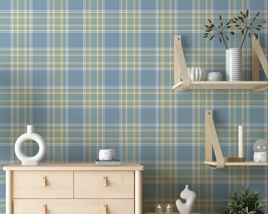 A bright bedroom or living area featuring a light blue, sage green, and white plaid wallpaper accent wall. Centered against the wall is a light birch chest of drawers with simple leather tab pulls, holding a circular white ceramic vase and a book. To the left stands a large potted bird of paradise plant in a low, round grey concrete planter. To the right, a matching two-tiered wooden shelving unit with leather straps hangs on the wall over a small nightstand holding lit candles and an abstract sculpture.