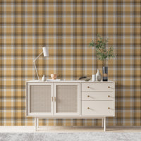 A closeup of a curated tabletop setting featuring a warm honey-gold plaid wallpaper with brown and cream accents. On the light-colored console surface, an assortment of earth-toned decor is arranged, including a small terracotta footed bowl on a white book to the left. To the right sits a collection of ribbed ceramic vases in white and cream, a shallow decorative bowl containing a sprig of dried pampas grass, and a tall, dark glass vase holding green leafy branches.