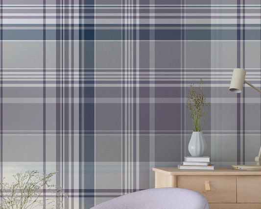 A stylish living room featuring a large plaid-patterned wallpaper in soft lilac, gray, and navy tones. The space includes a pale lavender accent chair, a round wooden side table with a small vase of flowers, a wooden cabinet with decor, and a tall green potted plant on dark wood flooring.