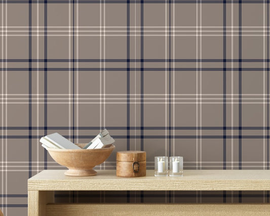 A cozy entryway setup is styled against a taupe, navy, and white plaid wallpaper background.
A long, light-colored wooden console table holds a bowl of books, a small round box, candles, and a white vase with delicate flowers.
Tucked neatly beneath the table are two woven storage baskets filled with magazines and reading materials.