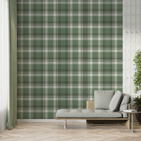 A modern living space featuring a prominent olive green and white plaid wallpaper accent wall. In front of the wall sits a sleek grey chaise lounge with matching plush pillows, accompanied by a small, round metal side table. To the left, a large window is dressed with sheer white curtains and long, sage green drapes over herringbone wood flooring. To the right, a textured white feature wall provides contrast, with a large potted green plant in a dark ceramic planter standing in the corner.