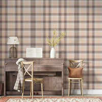 A closeup of a cozy interior setting featuring a dusty rose-pink and brown plaid wallpaper. On the left, a textured white ceramic vase with yellow flowering branches sits on top of a weathered, dark wood console table or upright piano. Next to it, a light wood cross-back dining chair holds a textured, rust-orange accent pillow.