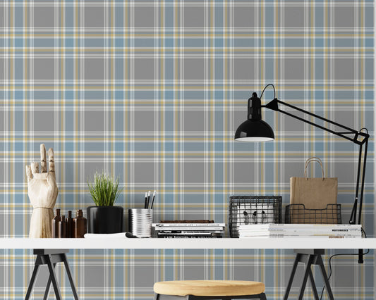A closeup of a modern desk workspace featuring a soft graphite grey plaid wallpaper with light blue and pale yellow accent lines. In the foreground, a white tabletop rests on black trestle legs, paired with a wooden stool. The desk is styled with a prominent black metal architect-style desk lamp on the right, stacks of design books and magazines, wire storage baskets, a small potted plant, a silver pencil cup, and an articulated wooden mannequin hand decoration on the far left.