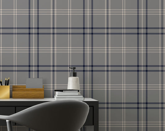 A minimalist home office workspace is set against a wall covered in grey, navy blue, and white plaid wallpaper.
In the center, a sleek black desk features a slim drawer built into its frame. Atop the desk sits a small table lamp with a grey base and a white cylindrical shade, a small square black photo frame, a gold pencil holder filled with pens, a wooden organizer, a stack of books, and a white soap dispenser bottle.A modern dark grey office chair with a curved backrest is pulled up to the desk, angled