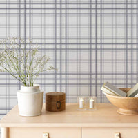 A serene indoor space features a soft stormy lavender and white plaid wallpaper.
The detailed design shows thin purple-grey lines intersecting to form clean grids across a light base.
In front, a light wooden tabletop holds books, a wooden bowl, and small candles.
The calming setup is beautifully accented with a white vase containing delicate white flowers.