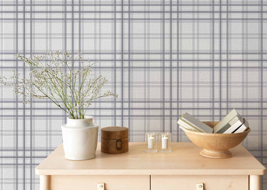 A serene indoor space features a soft stormy lavender and white plaid wallpaper.
The detailed design shows thin purple-grey lines intersecting to form clean grids across a light base.
In front, a light wooden tabletop holds books, a wooden bowl, and small candles.
The calming setup is beautifully accented with a white vase containing delicate white flowers.