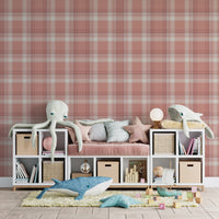 A children's playroom or nursery featuring a dusty terracotta pink and white plaid wallpaper accent wall. A long white storage unit sits against the wall, incorporating cubbies filled with books, storage baskets, and a central built-in bench with a pink velvet cushion and star-shaped pillows. Large plush sea creature toys, including a white octopus, a grey whale, and a blue whale, are scattered across the storage unit and on a yellow textured rug below. The room is finished with light grey wood plank floori