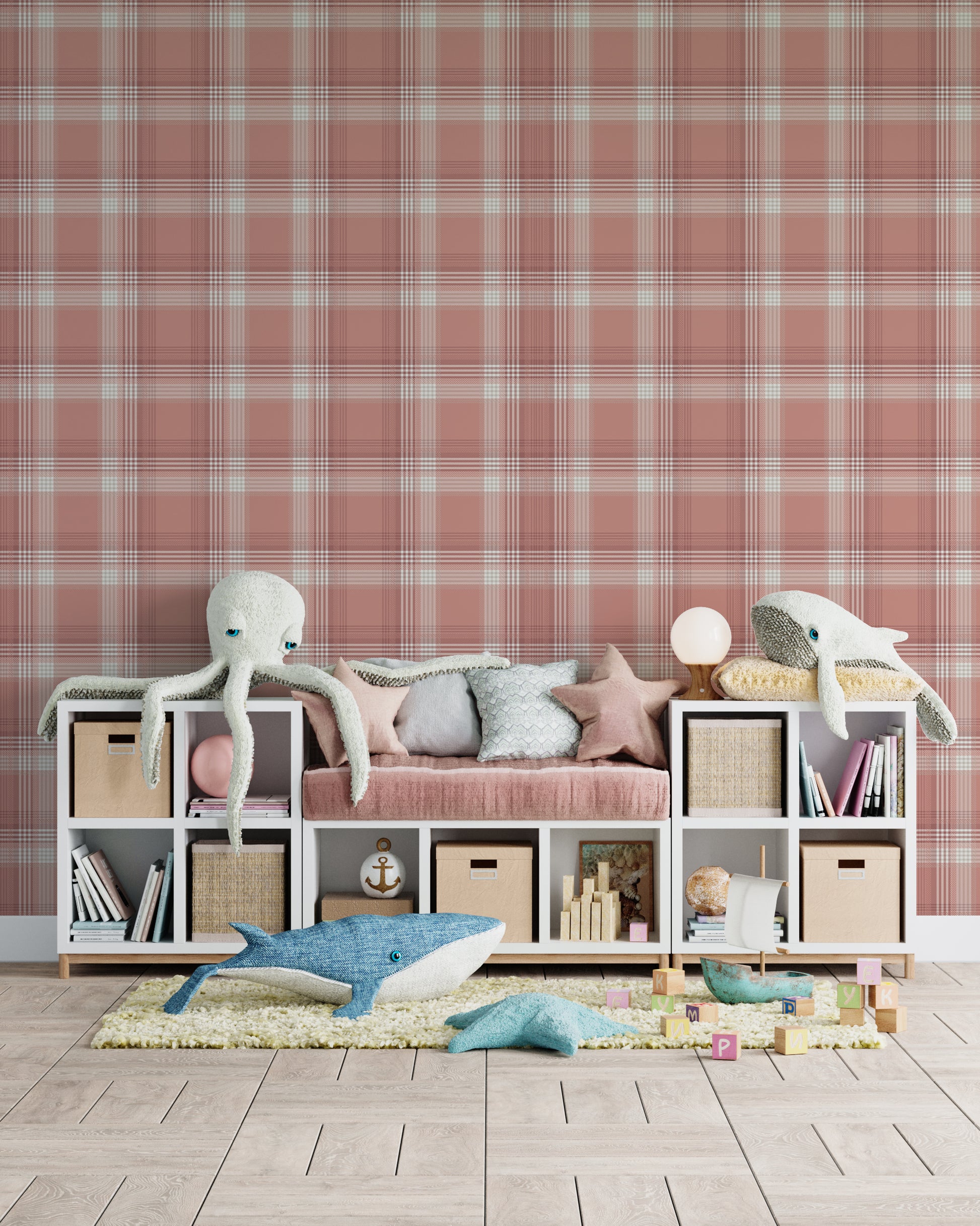 A children's playroom or nursery featuring a dusty terracotta pink and white plaid wallpaper accent wall. A long white storage unit sits against the wall, incorporating cubbies filled with books, storage baskets, and a central built-in bench with a pink velvet cushion and star-shaped pillows. Large plush sea creature toys, including a white octopus, a grey whale, and a blue whale, are scattered across the storage unit and on a yellow textured rug below. The room is finished with light grey wood plank floori