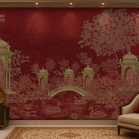 Elegant living room featuring a heritage-inspired mural in rich crimson red and antique gold. The mural showcases ornate domed pavilions, lush botanical foliage, an arched bridge, blooming lotus flowers, and graceful swans on a tranquil pond, with subtle palace architecture in the background. The space is styled with a cream upholstered accent chair, a burgundy embroidered cushion, dark wood furniture, brass decorative accents, a patterned beige rug, and warm ambient lighting, creating a luxurious atmospher