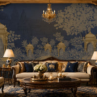 Luxurious living room featuring a royal Indian-inspired interior with a large navy blue and antique gold mural depicting domed pavilions, lush trees, and intricate architectural motifs. The space includes an ivory upholstered sofa, matching armchairs, carved dark wood furniture, navy and gold accent cushions, warm table lamps, a crystal chandelier, a round wooden coffee table with candles and flowers, and an ornate blue patterned rug, creating an elegant and opulent atmosphere.