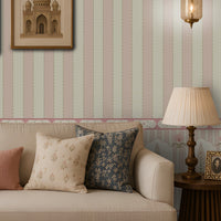 Elegant living room with a cream sofa and patterned blue, cream, and dusty-pink cushions, a carved wooden coffee table, cane armchair, decorative side tables, framed botanical and architectural artwork, brass lanterns, and pastel striped floral wallpaper.
