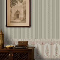 A styled interior scene showing a wall with vertical striped wallpaper in muted sage green, blush pink, and cream, plus a decorative border. A dark wood cabinet sits along the bottom with a brass lantern, a bowl of gold coins, stacked books, and a framed sepia architectural print hanging above.