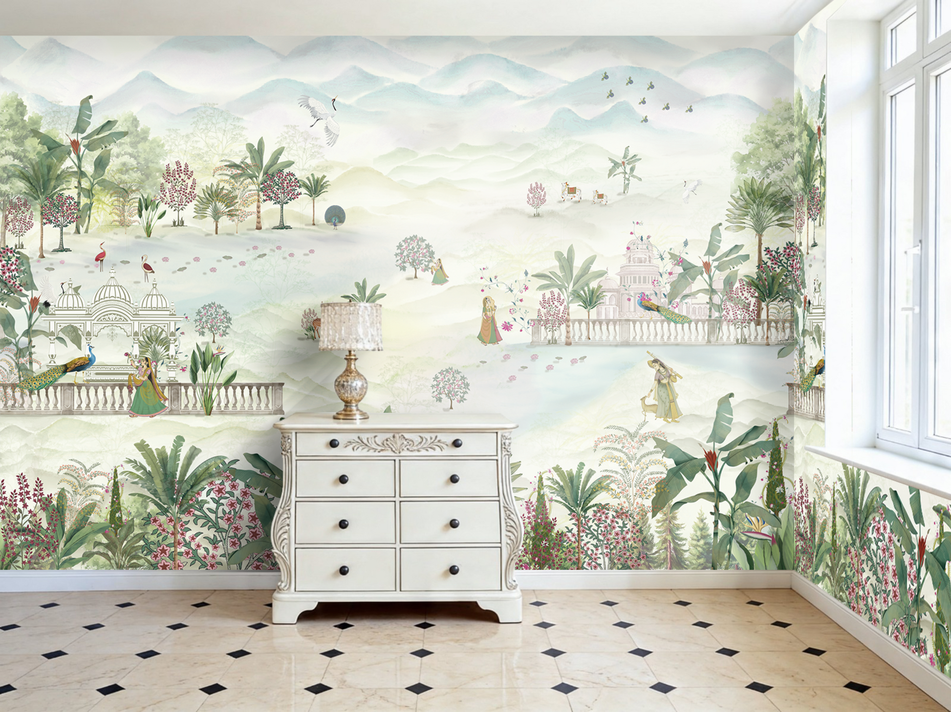 Bright, airy room with a large panoramic wallpaper mural depicting a dreamy pastel landscape. The mural features rolling misty mountains, tropical plants, flowering trees, peacocks, flamingos, cranes, and small figures in ornate gardens and palace settings. A white vintage-style dresser with a decorative lamp sits against the wall, while tall windows on the right let in natural light. The floor is tiled in cream marble with small black diamond accents