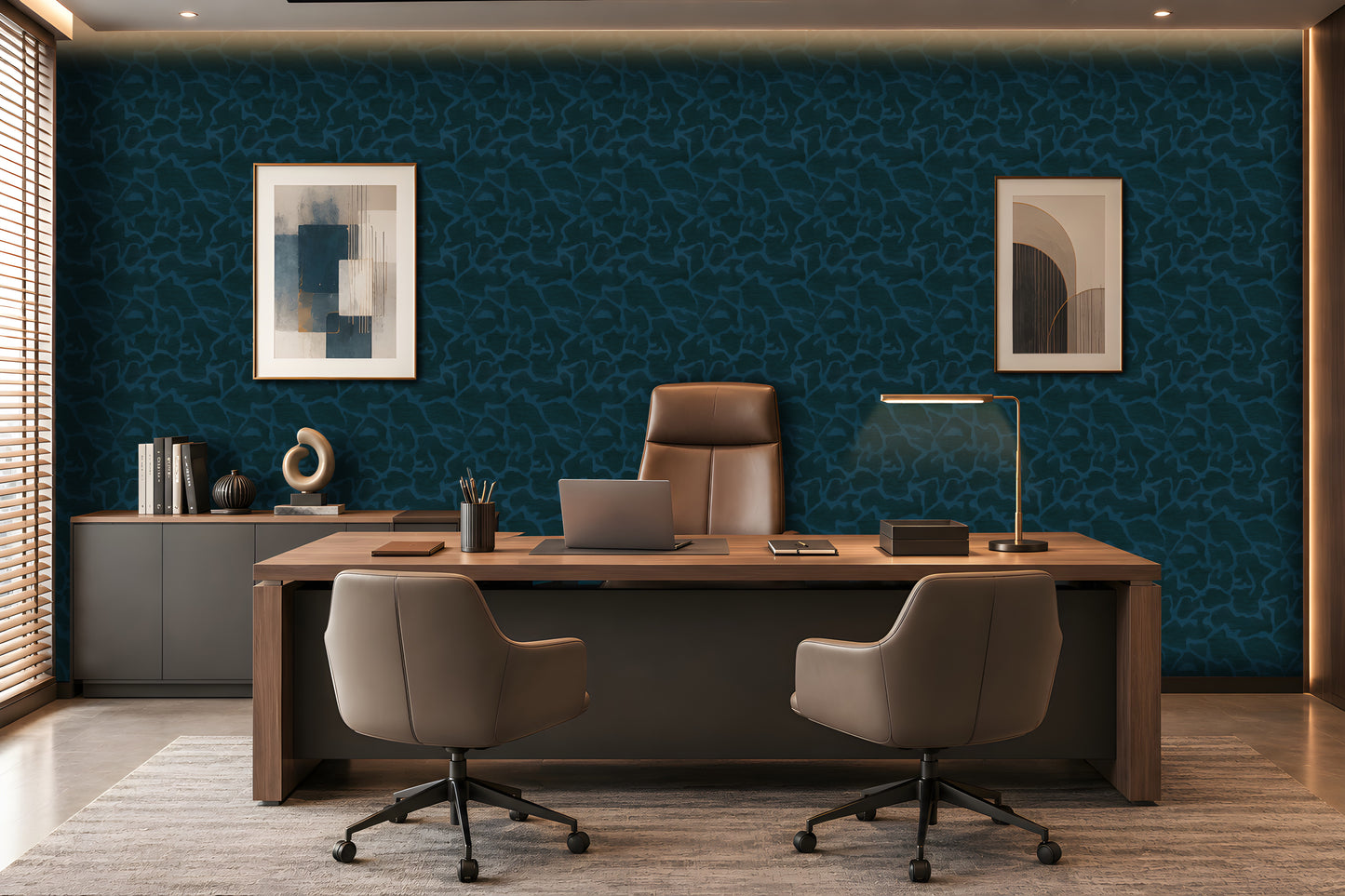 A modern office setup featuring a large wooden desk with two gray swivel chairs, a laptop, and a desk lamp, set against a dark blue patterned wallpaper wall with framed art.
