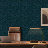 Dark teal patterned wallpaper in an office setting behind a wooden desk, a brown leather chair, a laptop, and a modern desk lamp, with a framed abstract print on the wall.